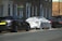 A street scene featuring several parked cars. One car is covered in white foam, suggesting it is undergoing a wash or cleaning. The background includes a brick residential building with several windows and doorways. A 'sold' sign with a phone number is visible near the building.