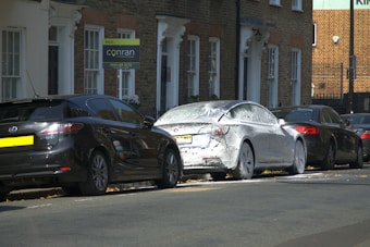 A street scene featuring several parked cars. One car is covered in white foam, suggesting it is undergoing a wash or cleaning. The background includes a brick residential building with several windows and doorways. A 'sold' sign with a phone number is visible near the building.