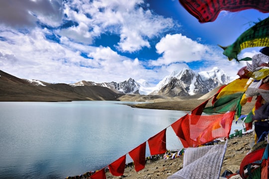 assorted flags on beach shore near snow covered mountains under blue and white sunny cloudy sky in Gurudongmar Lake India