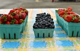 On a colorful tablecloth featuring floral patterns, several teal cartons of fresh fruits are organized. The cartons on both sides are filled with ripe red strawberries, while the central carton contains plump blueberries. The scene evokes a sense of freshness and abundance, typical of a farmer's market display.