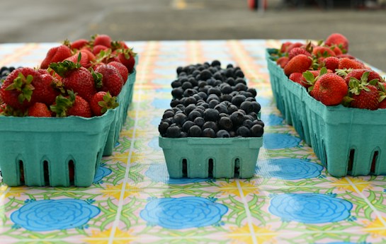 On a colorful tablecloth featuring floral patterns, several teal cartons of fresh fruits are organized. The cartons on both sides are filled with ripe red strawberries, while the central carton contains plump blueberries. The scene evokes a sense of freshness and abundance, typical of a farmer's market display.