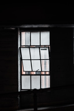 A dimly lit industrial-style window with a grid pattern is partially open. The glass has a textured appearance, possibly wire-reinforced, and casts a muted light into a shadowy interior space.