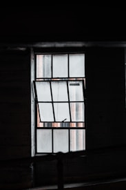 A dimly lit industrial-style window with a grid pattern is partially open. The glass has a textured appearance, possibly wire-reinforced, and casts a muted light into a shadowy interior space.