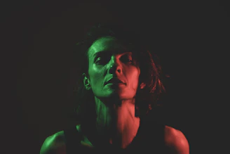 A person is partially illuminated by dramatic lighting, featuring green and red hues on their face and shoulders. The contrasting colors create a moody and intense atmosphere with deep shadows obscuring part of the face.