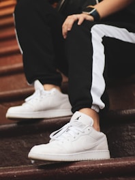 A person is sitting on stairs wearing black and white track pants and white athletic sneakers. The focus is on the shoes, which are positioned on the edge of a dark brown step. The person is leaning forward slightly, with one hand resting on their knee.