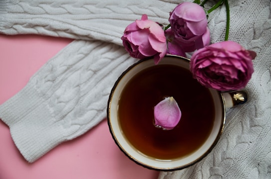 A cozy arrangement featuring a cup of tea placed on a soft, white cable-knit sweater. Pink roses are positioned around the cup, which has a single pink petal floating on the tea. The background is a soft pink color, enhancing the gentle and warm aesthetic.