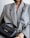 A person wearing a large, textured gray blazer holds a small black leather handbag with gold lettering and hardware. The handbag features a handle and an adjustable strap. The person is also wearing a thick, gold twisted bracelet.