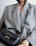 A person wearing a large, textured gray blazer holds a small black leather handbag with gold lettering and hardware. The handbag features a handle and an adjustable strap. The person is also wearing a thick, gold twisted bracelet.