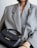 A person wearing a large, textured gray blazer holds a small black leather handbag with gold lettering and hardware. The handbag features a handle and an adjustable strap. The person is also wearing a thick, gold twisted bracelet.