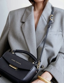 A person wearing a large, textured gray blazer holds a small black leather handbag with gold lettering and hardware. The handbag features a handle and an adjustable strap. The person is also wearing a thick, gold twisted bracelet.