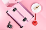 A pink metallic makeup case is surrounded by various cosmetic products. The items include a round makeup powder compact, a foundation bottle placed on a white plate, a red lipstick, and a black mascara. The background is soft pink, creating a cohesive and feminine aesthetic.