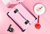 A pink metallic makeup case is surrounded by various cosmetic products. The items include a round makeup powder compact, a foundation bottle placed on a white plate, a red lipstick, and a black mascara. The background is soft pink, creating a cohesive and feminine aesthetic.