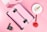 A pink metallic makeup case is surrounded by various cosmetic products. The items include a round makeup powder compact, a foundation bottle placed on a white plate, a red lipstick, and a black mascara. The background is soft pink, creating a cohesive and feminine aesthetic.