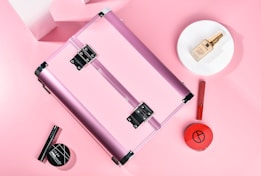 A pink metallic makeup case is surrounded by various cosmetic products. The items include a round makeup powder compact, a foundation bottle placed on a white plate, a red lipstick, and a black mascara. The background is soft pink, creating a cohesive and feminine aesthetic.
