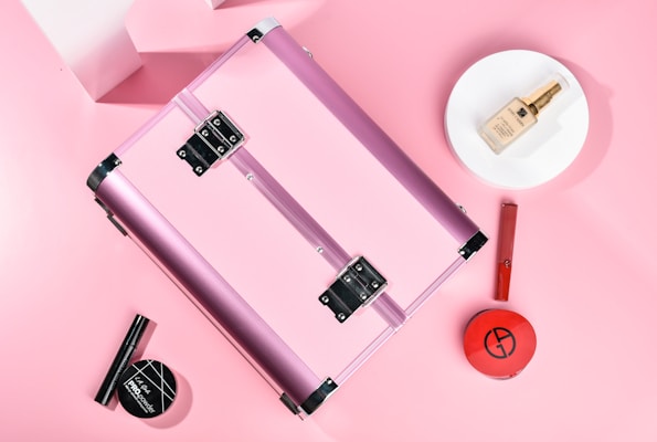 A pink metallic makeup case is surrounded by various cosmetic products. The items include a round makeup powder compact, a foundation bottle placed on a white plate, a red lipstick, and a black mascara. The background is soft pink, creating a cohesive and feminine aesthetic.