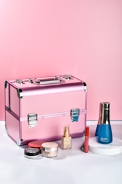 A pink makeup case is placed on a surface surrounded by various cosmetic products, including a blue bottle, a foundation bottle, a red lipstick, and multiple small containers of makeup powders.