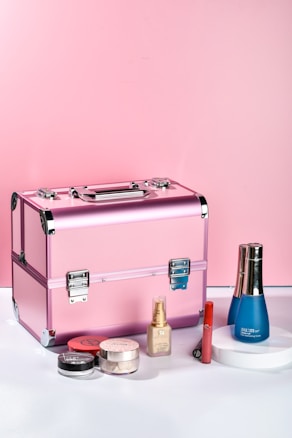A pink makeup case is placed on a surface surrounded by various cosmetic products, including a blue bottle, a foundation bottle, a red lipstick, and multiple small containers of makeup powders.