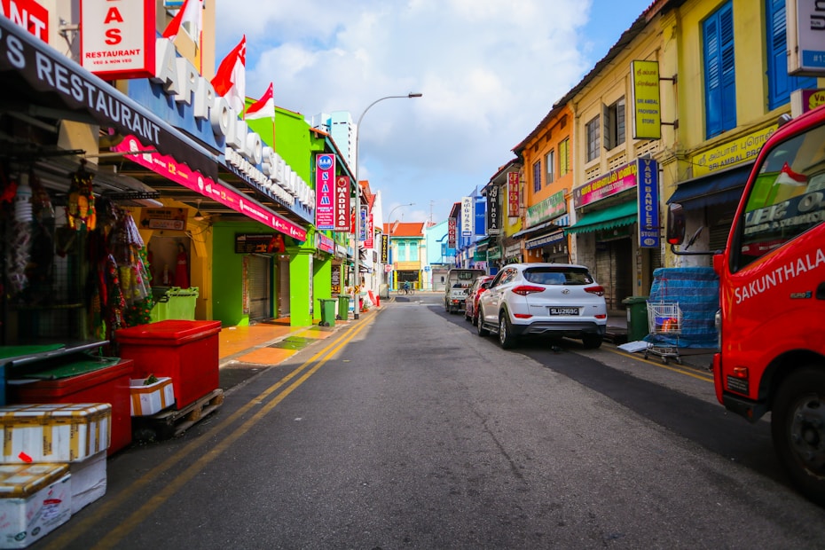 Colorful storefronts and buildings line both sides of a narrow street. Cars are parked along the road, and signs with various colors and languages are visible. The atmosphere is vibrant and busy, with red, green, and yellow facades contributing to the lively scene. Pedestrian pathways and various shop signs add to the urban environment.