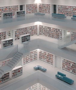 A modern, multi-story library interior with white walls and shelves filled with books. The design is open with balconies and staircases providing access to multiple levels. Minimalist furniture, such as light blue benches, is arranged neatly on each floor.