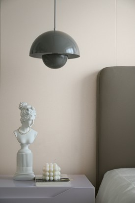 A modern bedroom setting featuring a minimalist design. A dark grey pendant lamp hangs from the ceiling above a bedside table. The table holds a white marble bust adorned with a pearl necklace, next to a decorative candle resembling a cluster of bubbles. An upholstered headboard is partially visible against a light-colored wall.