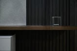 A minimalist interior featuring a wooden shelf against a dark, vertically paneled wall. A set of Newton's Cradle balance balls is placed on the shelf. The left side shows a small white cabinet partially visible under the shelf.