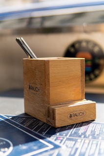 A wooden pencil holder with two pens inside is placed on a surface that appears to have blueprints or technical drawings. The pencil holder has the word 'RAUCH' printed on it. In the background, there's a blurred circular object with colorful elements.