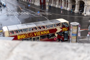 A double-decker bus with 'Big Bus Vienna' written on it is parked on a wet city street. The bus is red and cream in color, with an open top and empty seats visible. Nearby, there are pedestrians taking photos and signs advertising local exhibitions. The surroundings are urban, with old stone buildings and some traffic signage.