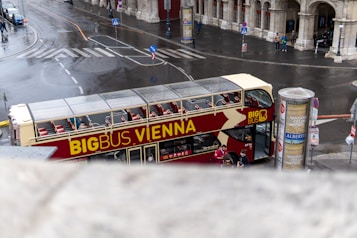 A double-decker bus with 'Big Bus Vienna' written on it is parked on a wet city street. The bus is red and cream in color, with an open top and empty seats visible. Nearby, there are pedestrians taking photos and signs advertising local exhibitions. The surroundings are urban, with old stone buildings and some traffic signage.