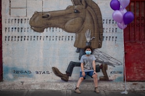 A child wearing a face mask sits in front of a mural depicting a surreal horse with human-like legs and wings, painted on a brick wall. The child is holding a bunch of purple balloons and is dressed casually. The mural is signed with the names Pegas Stas and Kislow.