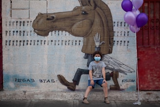 A child wearing a face mask sits in front of a mural depicting a surreal horse with human-like legs and wings, painted on a brick wall. The child is holding a bunch of purple balloons and is dressed casually. The mural is signed with the names Pegas Stas and Kislow.