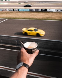 A person wearing a wristwatch holds a disposable coffee cup in the foreground, with a racing track in the background. A bright yellow sports car speeds past on the track, closely aligned with other distant vehicles. Advertising banners line the fence along the racetrack.