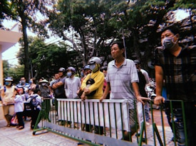 A group of people standing behind a green barrier, some wearing helmets and masks, are gathered under a canopy of trees. A few children in school uniforms are present in the foreground, suggesting a setting near a school or public institution.
