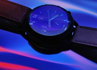 A wristwatch with a leather strap positioned against a vibrant background of blue and red hues. The watch display features visible hand markers and detailed watch face elements, such as dials and numeric indicators. The lighting creates a striking visual effect that highlights the watch's glass surface and metallic accents.