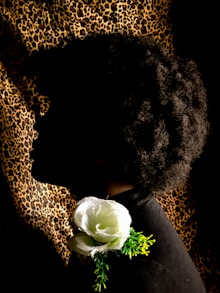 A silhouette of a person with an afro hairstyle set against a leopard print background. A large, white artificial flower is positioned near the neckline, with green foliage accenting it.