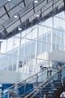 An interior architectural scene featuring large glass windows and a high ceiling with exposed beams. The staircase with metal and glass railings leads upwards. The scene is well-lit with modern ceiling lights and has a minimalist design.