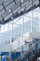 An interior architectural scene featuring large glass windows and a high ceiling with exposed beams. The staircase with metal and glass railings leads upwards. The scene is well-lit with modern ceiling lights and has a minimalist design.