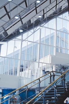 An interior architectural scene featuring large glass windows and a high ceiling with exposed beams. The staircase with metal and glass railings leads upwards. The scene is well-lit with modern ceiling lights and has a minimalist design.