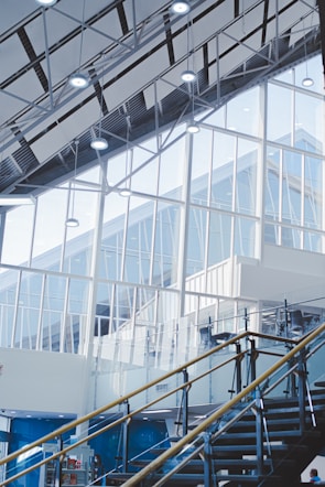 An interior architectural scene featuring large glass windows and a high ceiling with exposed beams. The staircase with metal and glass railings leads upwards. The scene is well-lit with modern ceiling lights and has a minimalist design.