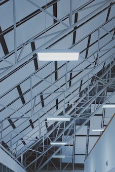 An architectural ceiling design featuring geometric patterns formed by metal beams and panels. Multiple fluorescent lights are suspended from the structure, creating an industrial and modern atmosphere.