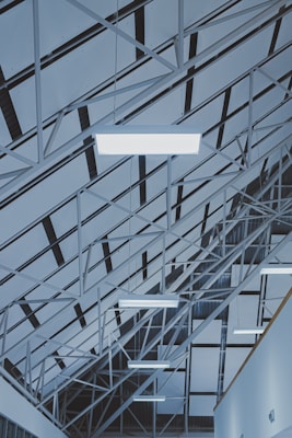 An architectural ceiling design featuring geometric patterns formed by metal beams and panels. Multiple fluorescent lights are suspended from the structure, creating an industrial and modern atmosphere.