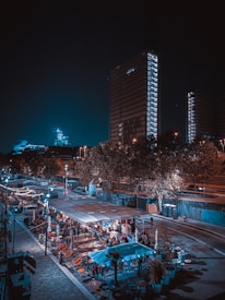 An urban nighttime scene featuring tall, illuminated buildings and a lively outdoor area with seating and string lights. People are gathered in an open-air setting, likely a restaurant or market, surrounded by trees and streetlights.