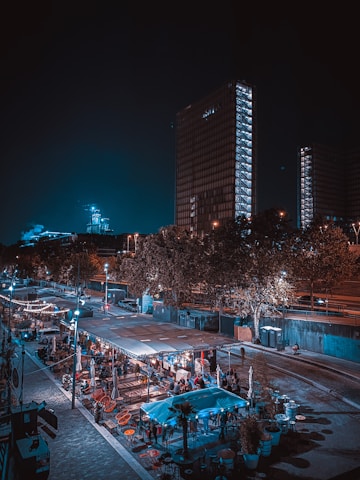 An urban nighttime scene featuring tall, illuminated buildings and a lively outdoor area with seating and string lights. People are gathered in an open-air setting, likely a restaurant or market, surrounded by trees and streetlights.
