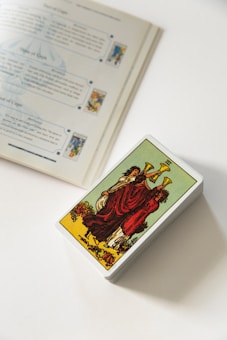 A tarot card deck is placed on a white surface next to an open book with descriptions and illustrations of various tarot cards. The top card on the deck displays the Three of Cups card depicting three women in red robes holding golden cups.