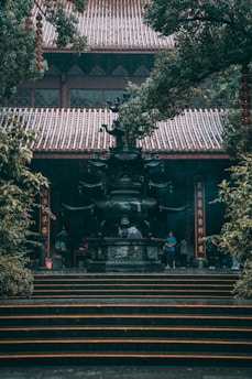 An ornate, large incense burner stands prominently on a series of stone steps, surrounded by lush green foliage. A traditional building with a tiled roof serves as the backdrop, with lanterns and Chinese calligraphy decorating the scene. Several people are gathered near the burner, creating a sense of activity.