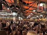 A large indoor gathering with people dressed in traditional Bavarian clothing, enjoying a festive atmosphere. The space is decorated with green garlands and colorful flags, and the lighting casts a warm glow. The crowd is dense, with many people holding drinks and engaging in conversation.