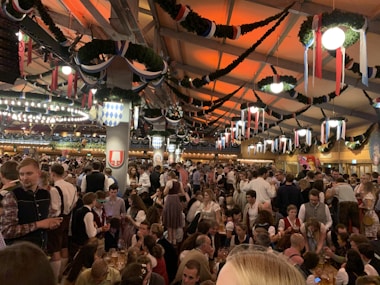 A large indoor gathering with people dressed in traditional Bavarian clothing, enjoying a festive atmosphere. The space is decorated with green garlands and colorful flags, and the lighting casts a warm glow. The crowd is dense, with many people holding drinks and engaging in conversation.