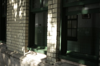 Exterior view of a building with green window frames and light brick walls. Shadows from nearby trees create a dappled effect on the facade. Part of the interior, featuring green tiled walls, can be seen through the windows.