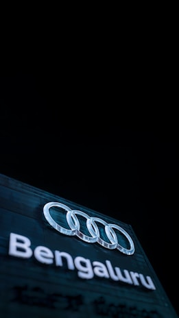 An illuminated sign displaying the Audi logo above the word 'Bengaluru', set against a dark background. The logo consists of four interlocking rings, which are prominently lit.