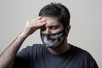 A person wearing a camouflage-patterned face mask holds their forehead with their eyes closed, indicating a headache or discomfort. The person is dressed in a dark t-shirt with a neutral background.