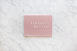 A pink pegboard with the words 'KINDNESS MATTERS' in white letters placed on a hexagonal tiled surface with a marble pattern.